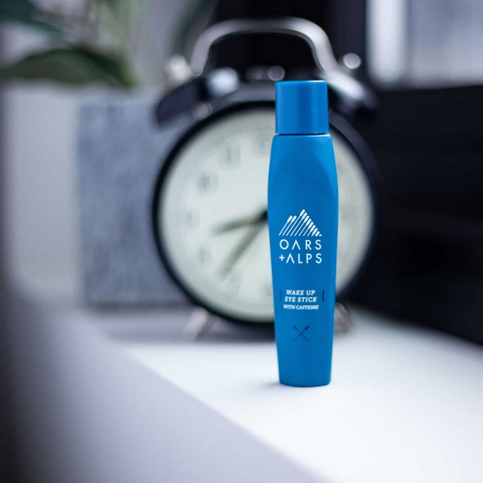 A blue Oars + Alps Wake Up Eye Stick sits on a white surface before a blurred clock, ideal for reducing puffiness and refreshing the under-eye area.