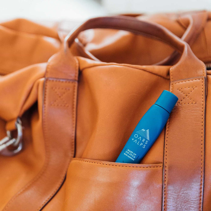 A blue Oars + Alps Wake Up Eye Stick is tucked into the front pocket of a light brown leather travel bag.
