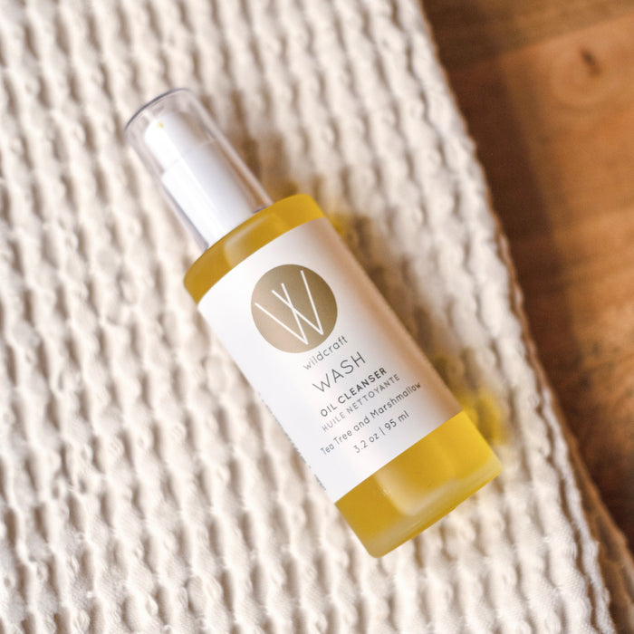 A bottle of Wildcraft Wash Oil Cleanser sits on a textured white cloth, featuring a sleek design with a white and gold label showcasing the brands logo and details. Infused with botanical oils like turmeric and marshmallow root, it pairs beautifully with the wooden backdrop.