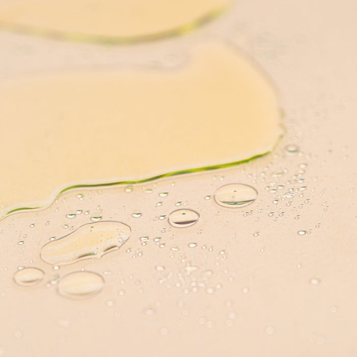 A close-up of clear, oily droplets and smears from Wildcrafts Wash Oil Cleanser reveals an amber sheen. Small bubbles and botanical oils create a glossy texture against the light surface.