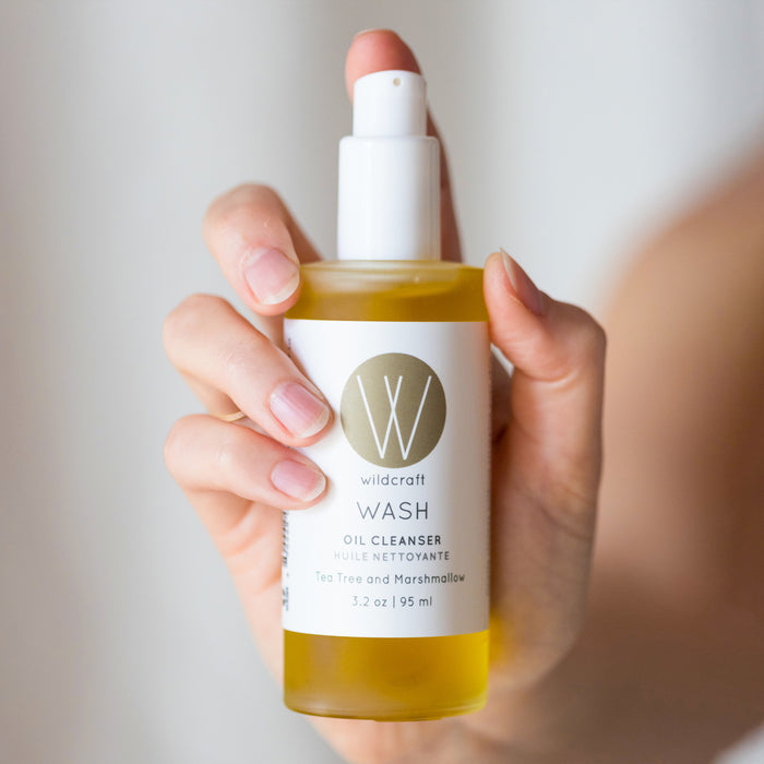 A hand cradles Wildcrafts Wash Oil Cleanser, infused with tea tree and marshmallow oils. The clear 3.2 oz (95 ml) bottle has a white label, gold circle logo, and a white pump dispenser for convenience.