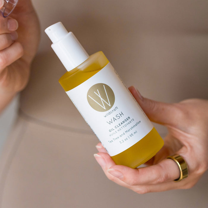 A person holds a yellow bottle of Wildcrafts Wash Oil Cleanser, enriched with botanical oils. The bottle features a white label with the brand logo and product details. With a gold ring adorning their hand, they grip the clear cap, highlighting the elegance of this cleanser.