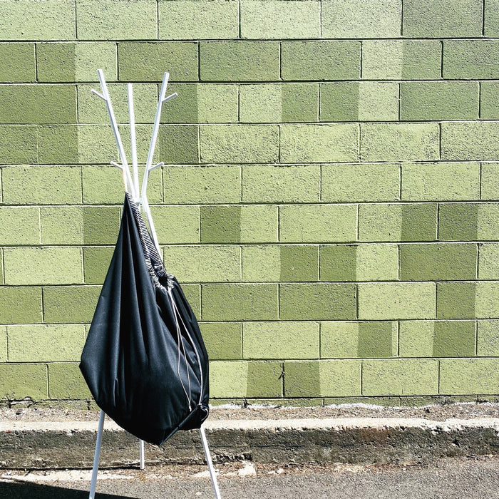 A white metal coat rack stands on pavement against a green block wall, with a large Washable Trash & Recycling Bag by Marleys Monsters, made of water-resistant fabric, hanging from it and casting a shadow on the wall. The wall behind showcases various shades of green in a pattern.