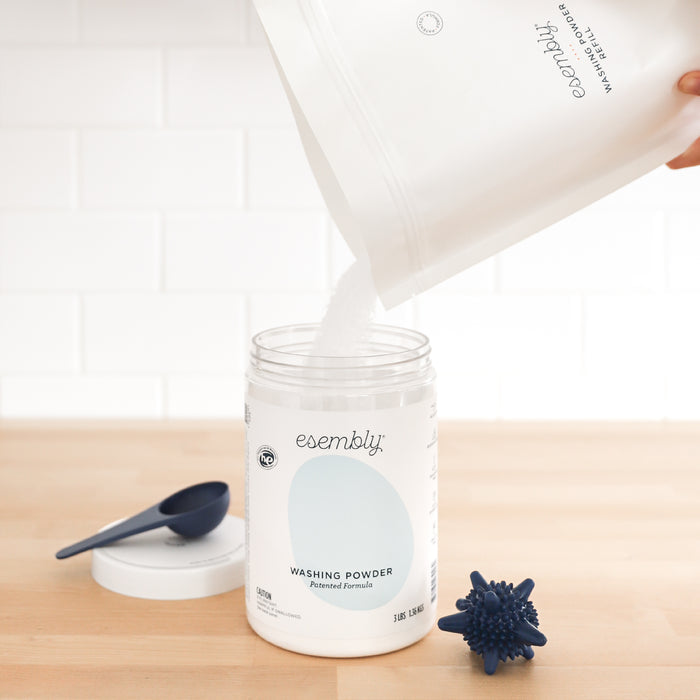 A person pours Esembly Baby Washing Powder from a white bag into an open jar labeled Esembly Washing Powder, with a lid and blue scoop nearby on a wooden surface against a white tiled wall, perfect for keeping cloth diapers fresh and clean.