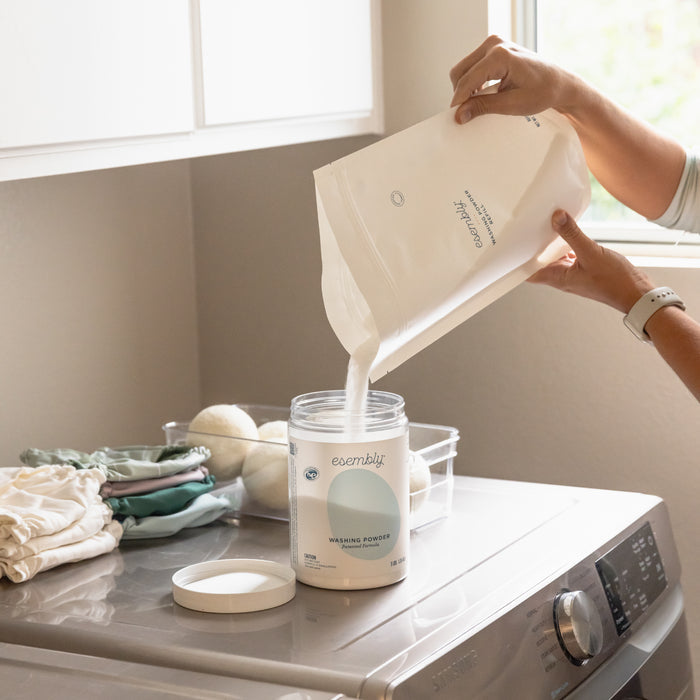 Someone pours Esembly Babys Washing Powder into a container over the washing machine. Nearby, eco-friendly cloth diapers and dryer balls are placed. Sunlight filters through a window, creating a soft, clean atmosphere.
