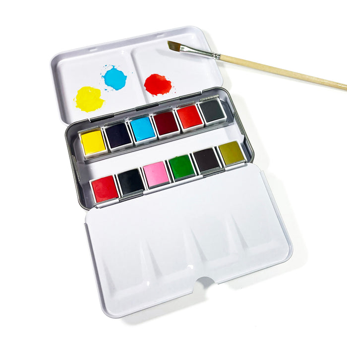 The Elseware Unplug Watercolor Set features twelve travel-size color pans, a mixing tray with blue and yellow spots, and a paintbrush atop an eco-friendly aluminum case, all presented on a white background.