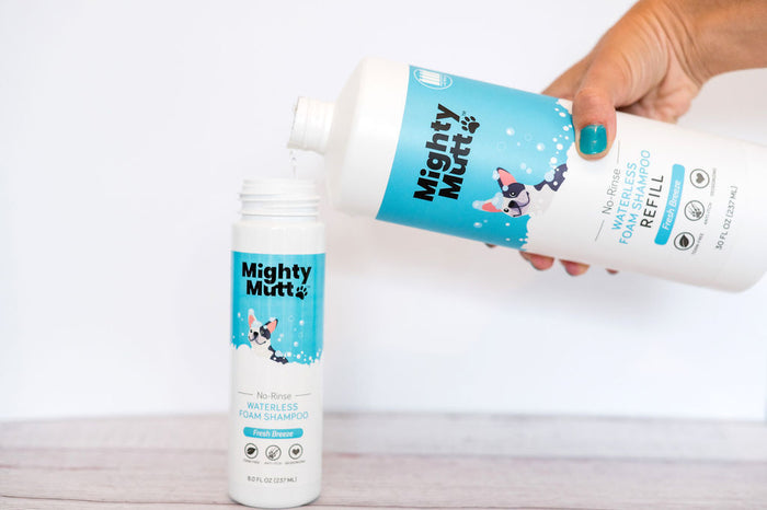 A hand transfers Mighty Mutts Waterless Foam Pet Shampoo Refill into a smaller bottle. The bottles, featuring a playful dog illustration, offer a hypoallergenic formula for no-rinse pet grooming against a light background.