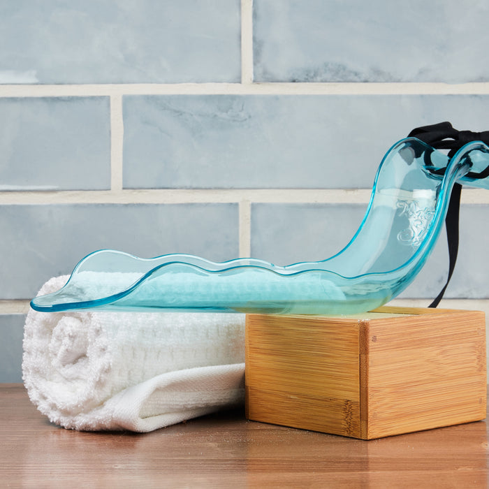 A glass sculpture rests on a wooden block beside a rolled white towel, subtly echoing Lovabilitys WaterSlyde design. Light blue tiles with white grout in the background enhance the serene ambiance.