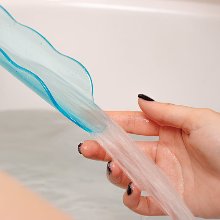 A hand with black nail polish holds a light blue, curved Lovability WaterSlyde, directing water over its edge. The scene reveals a filled bathtub with shimmering reflections—the perfect backdrop for an aquatic stimulator experience.