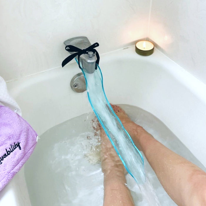 Someone relaxes in a bathtub, water flowing from the faucet through a blue Lovability WaterSlyde. A lit candle rests on the tubs edge, and a purple towel with text enhances this peaceful retreat.