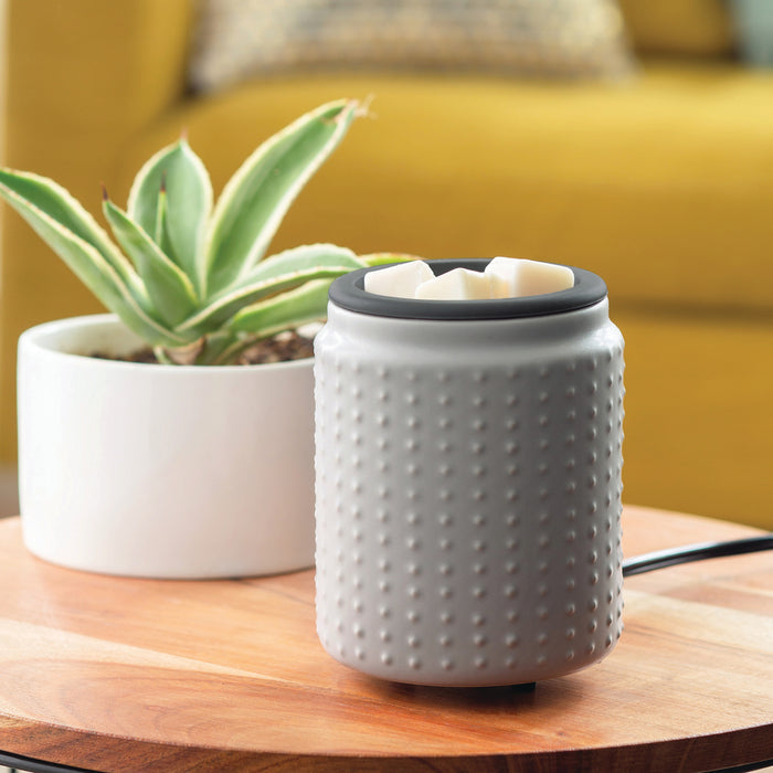 A Candle Warmers Etc. Wax Warmer with a silicone dish, featuring a textured design, is on a wooden table beside a white pot with a green succulent. A yellow couch is in the background, and its plugged-in state suggests its in use.