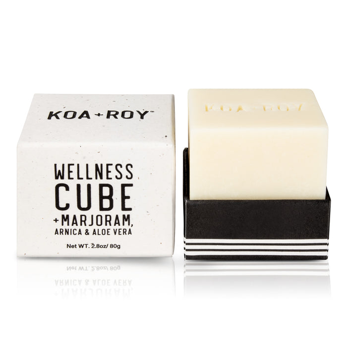 A KOA + ROY Wellness Cube is showcased, featuring all-natural skincare benefits. The white cube-shaped soap lies in a black holder next to its box, which lists ingredients like marjoram, arnica, and aloe vera. At 2.8 oz (80g), its ideal for an aromatherapy experience.