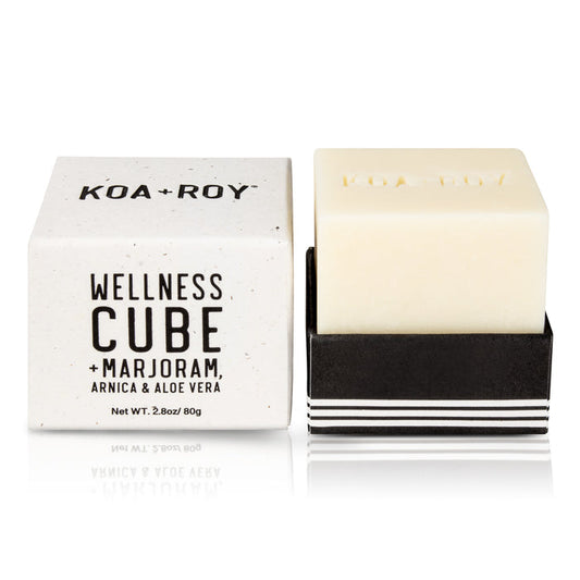 A KOA + ROY Wellness Cube is showcased, featuring all-natural skincare benefits. The white cube-shaped soap lies in a black holder next to its box, which lists ingredients like marjoram, arnica, and aloe vera. At 2.8 oz (80g), its ideal for an aromatherapy experience.