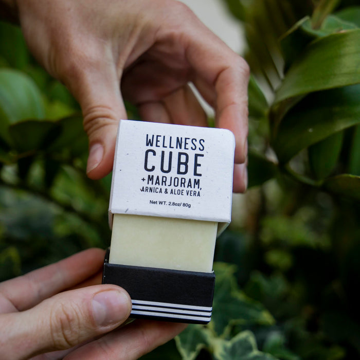 Someone holds a KOA + ROY Wellness Cube box containing an all-natural skincare bar of soap slightly slid out. The setting, decorated with green leafy plants, creates a calming ambiance ideal for aromatherapy.