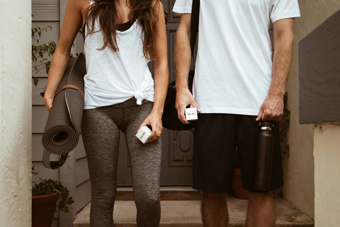 Outside the KOA + ROY Wellness Cube, a woman in a white tank top and gray leggings and a man in a white T-shirt and black shorts hold coffee cups, rolled yoga mats, and water bottles as they discuss their favorite aromatherapy blends.