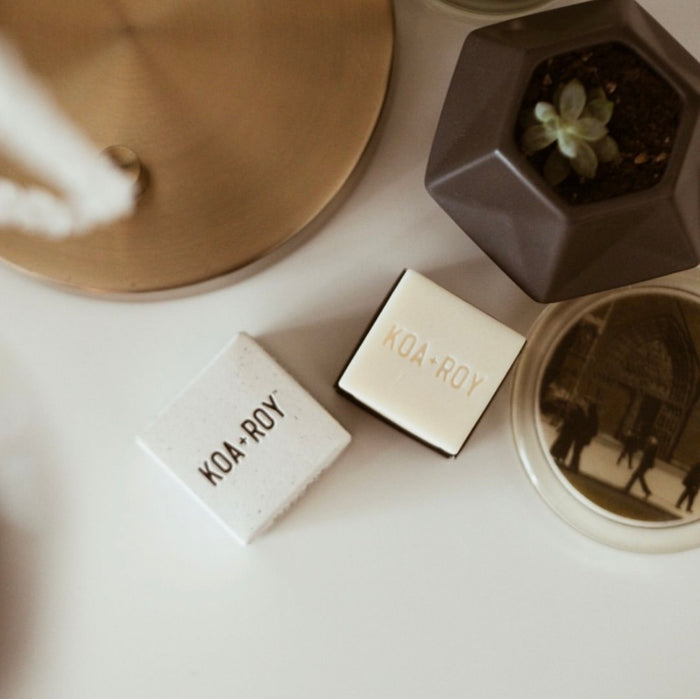 A tabletop features two KOA + ROY small boxes, a geometric planter with a succulent, and a gold circular item. A vintage photo coaster complements the setup, enhanced by the aromatherapy of a KOA + ROY Wellness Cube.