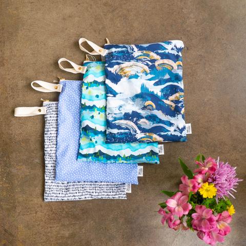 Five colorful Marleys Monsters Wet Bags with white handles are displayed on a brown surface, featuring wave, dot, and abstract patterns. A small bouquet of pink, purple, and yellow flowers complements the vibrant pouches.