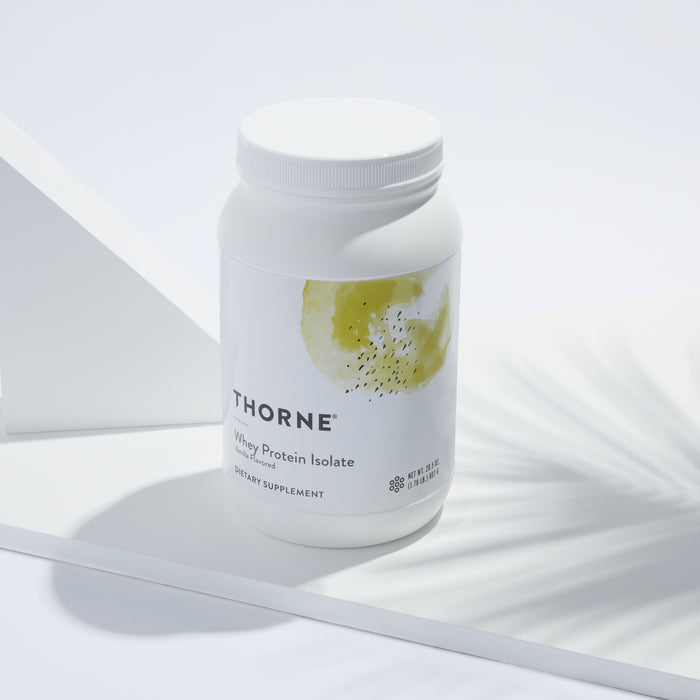The Whey Protein Isolate from Thorne, in a white container with abstract artwork, offers bioavailable protein for muscle repair. It rests elegantly on a white surface, complemented by soft shadows and geometric shapes for a clean, modern aesthetic.