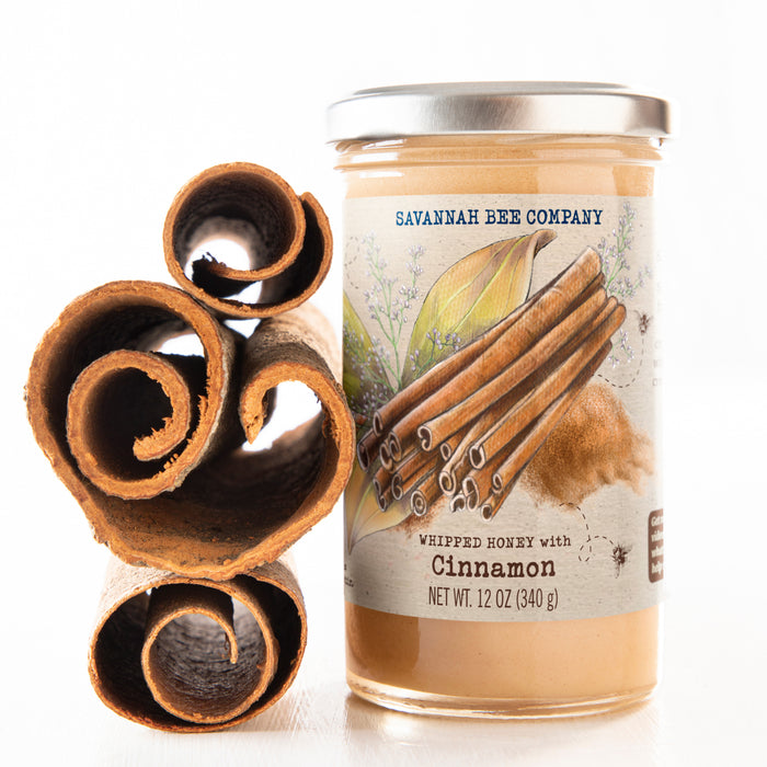 A jar of Savannah Bee Company Whipped Honey with Cinnamon sits beside cinnamon sticks, featuring a label with cinnamon and honeycomb illustrations, encapsulating its 12 oz (340 g) essence.