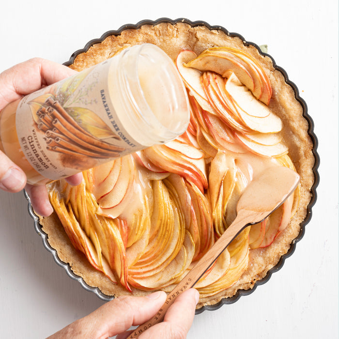 Hands pour Savannah Bee Companys Whipped Honey with Cinnamon over a freshly made apple tart in a round pan. The tart displays thinly sliced apples arranged in a spiral pattern, and a spatula helps spread the mixture evenly.