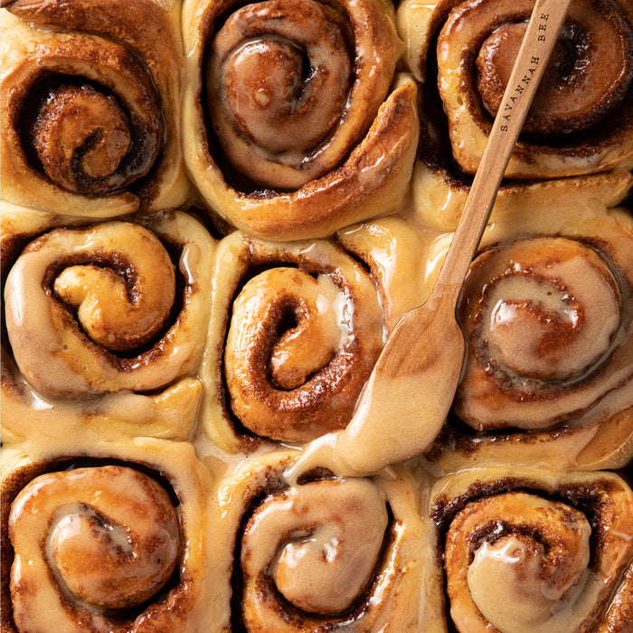 A close-up of nine glazed cinnamon rolls in a baking dish highlights one roll with golden-brown swirls and gooey filling as a wooden spoon drizzles Savannah Bee Companys Whipped Honey with Cinnamon, infusing a warm glaze over the tightly packed treats.