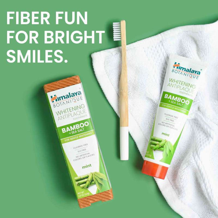 Toothpaste and toothbrush on a white towel with green background. The toothpaste, labeled "Himalaya Botanique Whitening Antiplaque," features bamboo and mint. A wooden toothbrush lies beside it. Text reads: "Fiber fun for bright smiles.