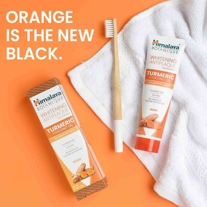 A packaging and tube of Himalaya Botanique Turmeric &amp; Coconut Oil Whitening Antiplaque toothpaste in mint flavor are displayed on an orange background. A beige-handled toothbrush and a white towel are nearby. Text reads, "Orange is the new black.