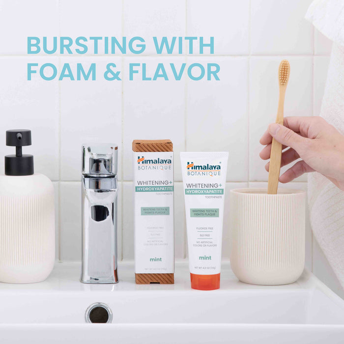 In a bathroom sink scene, a hand holds a toothbrush near a fluoride-free Himalaya Whitening+ Hydroxyapatite Toothpaste in mint flavor, next to a dispenser and faucet. Text reads Bursting with Foam & Flavor.