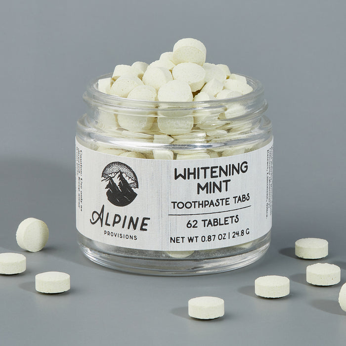 The clear jar of Alpine Provisions Whitening Mint Toothpaste Tabs, containing 62 small white tablets weighing 0.87 oz (24.8 g), is essential for healthy teeth and gums by preventing plaque buildup, with some tablets scattered on the surrounding gray surface.