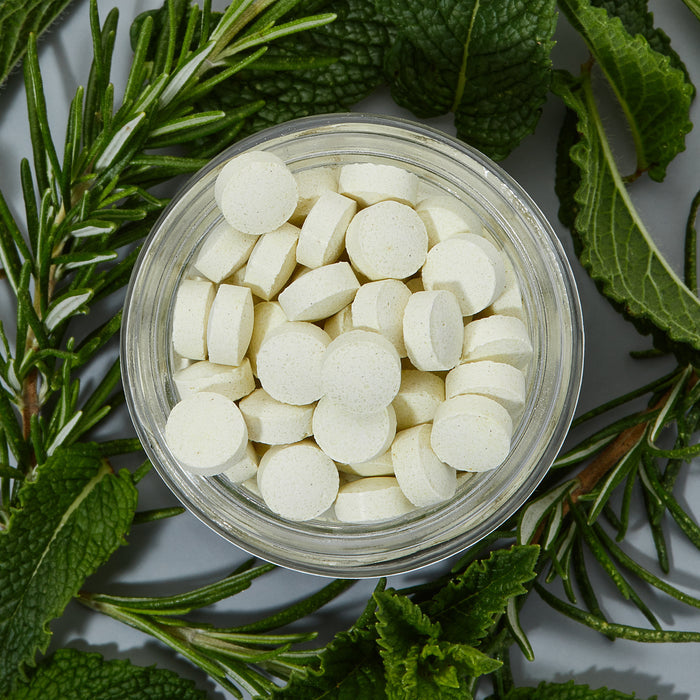 A glass jar of Alpine Provisions Whitening Mint Toothpaste Tabs sits on a gray surface surrounded by rosemary and mint, designed to promote healthy teeth and gums by preventing plaque buildup.