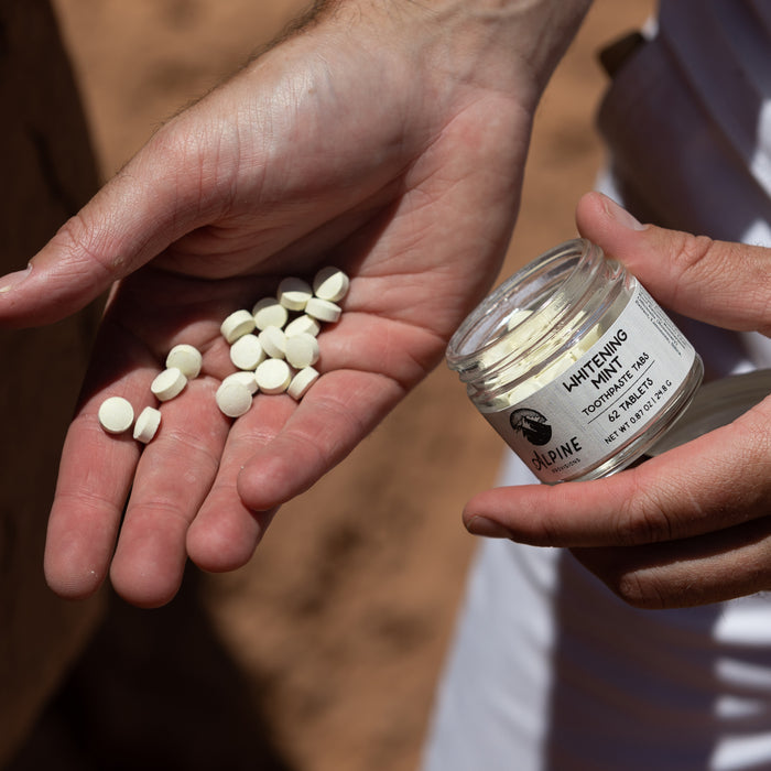 A person holds an open jar labeled Whitening Mint Toothpaste Tabs by Alpine Provisions, displaying round tabs in both hands. Designed to promote healthy teeth and gums, these tabs aid in preventing plaque buildup. A softly blurred background adds a gentle touch to the serene scene.