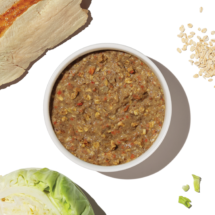 A bowl of The Honest Kitchen Dehydrated Whole Grain Turkey Dog Food sits on a white background, surrounded by raw chicken breast, oats, chopped cabbage, and green vegetables.