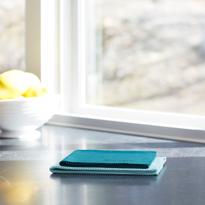 Teal microfiber cloths from the E-Cloth Window Cleaning Kit rest on a shiny countertop by a window. A white bowl with yellow lemons is slightly out of focus in the background. Natural light enhances the promise of streak-free window cleaning results.