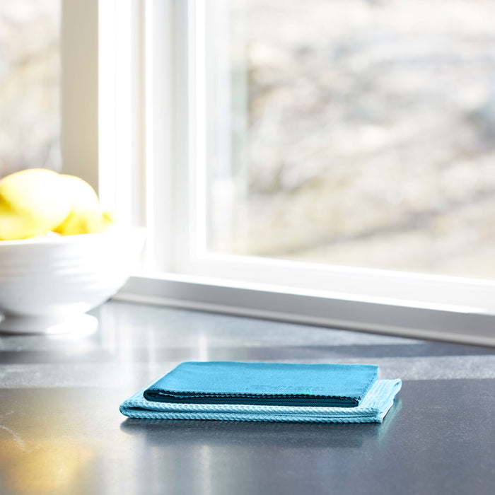 A stack of E-Cloth blue microfiber cloths is neatly folded on a shiny countertop. In the background, a blurred bowl of yellow apples sits next to a bright window, perfect for using the E-Cloth Window Cleaning Kit to achieve streak-free results in natural light.