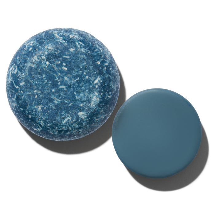 A blue, textured soap bar with white speckles sits on the left, infused with essential oils for sustainable self-care, next to a smooth blue disc. Both items are from the NOT!CE Hair Co Wingman Travel Set and cast shadows on a plain white background.