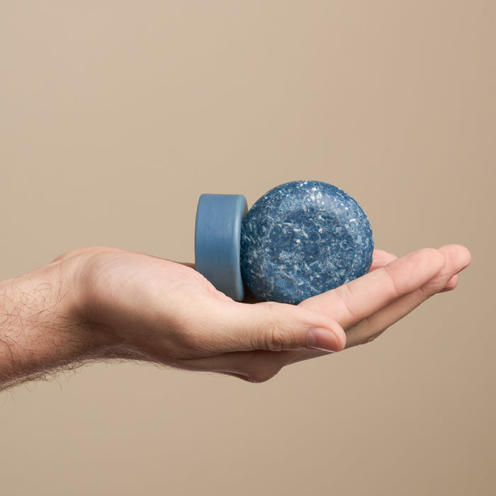 A hand holds the round blue NOT!CE Hair Co Wingman Travel Set soap bar and matching dish against a plain beige background, highlighting sustainable self-care with essential oils.