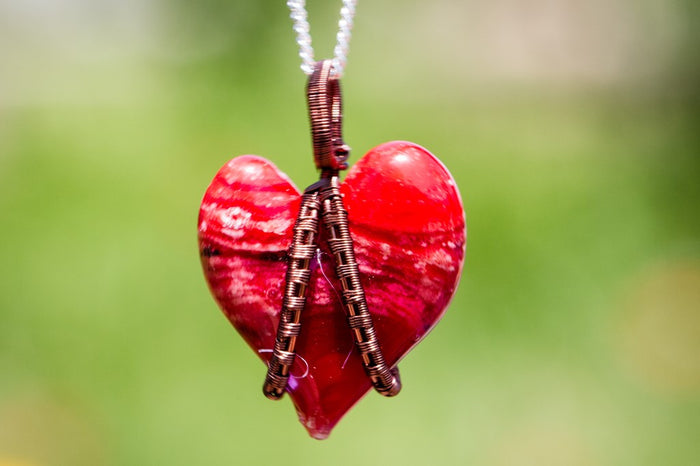 Red glass heart pendant wrapped in copper wire with a silver chain against a green background.