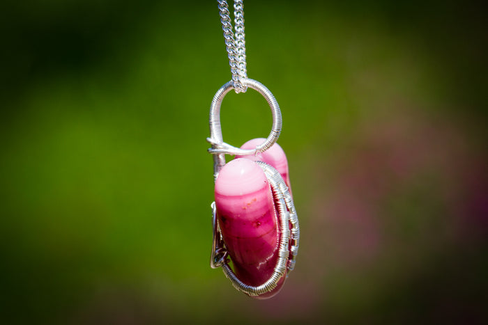 Pink glass heart pendant wrapped in silver wire with a matching chain against a blurred green backdrop.