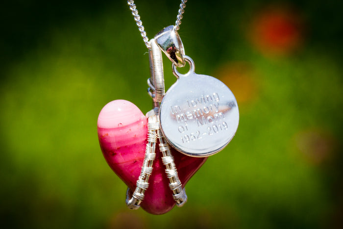 Pink glass heart cremation ashes pendant wrapped in silver wire on a silver chain against a blurred greenery background.