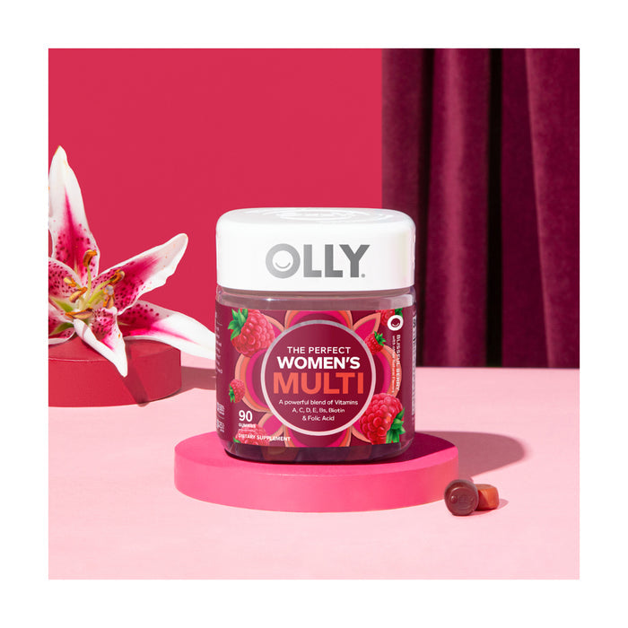 A jar of OLLY Womens Multi gummy vitamins, rich in B Vitamins, features pink and red packaging with raspberries. It sits beside a fresh pink lily against a soft pink and burgundy background, with a single gummy elegantly displayed in front.