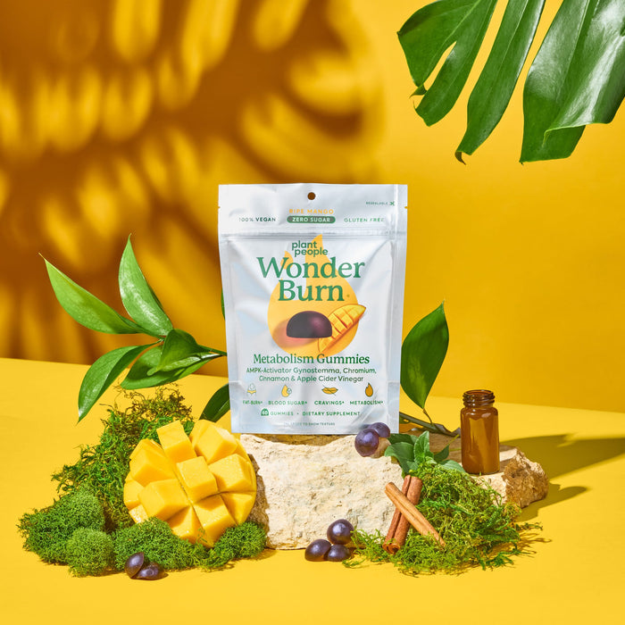 A bag of Plant People WonderBurn Gummies - For Metabolism & Cravings Control stands on a mossy rock, surrounded by cinnamon sticks, an amber bottle, mango, grapes, and tropical leaves with a yellow background.
