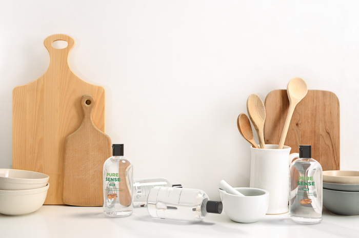 In a kitchen scene with wooden cutting boards and utensils against a white wall, three Pure Sense hand sanitizers sit among ceramic bowls and a mortar and pestle on the white countertop. Nearby, theres a bottle of Pure Sense Wood & Bamboo Cutting Board Conditioner, perfect for preserving woods luster.