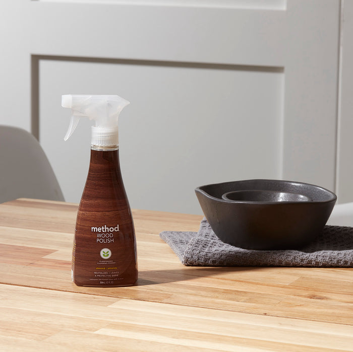 A bottle of method Wood Polish on a wooden table promises a natural shine, with two stacked black bowls on gray cloth beside it, set against a light wall. This sustainable choice exudes elegance and care in every detail.