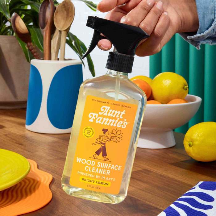 A hand holds Aunt Fannies Wood Surface Cleaner, featuring plant-based ingredients, on a kitchen counter with colorful dishes, fruit, and utensils in the background.