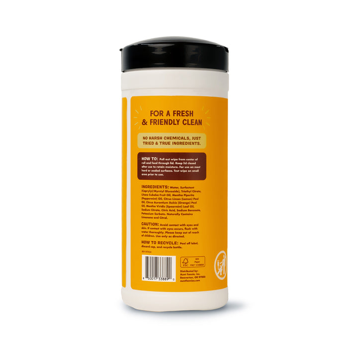 Aunt Fannies Wood Surface Wipes come in a cylindrical container with a yellow label and black lid, featuring usage instructions, cautionary notes, recycling info, and an ingredient list. The text highlights the natural formula using essential oils for cleaning.