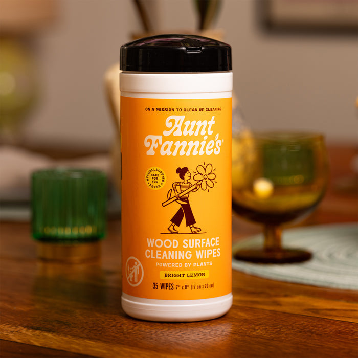 A bright yellow cylindrical container of Aunt Fannies Wood Surface Wipes sits on a table. The label, with cleaning graphic, features Powered by Plants, Bright Lemon, highlighting its natural formula. The background is softly blurred.
