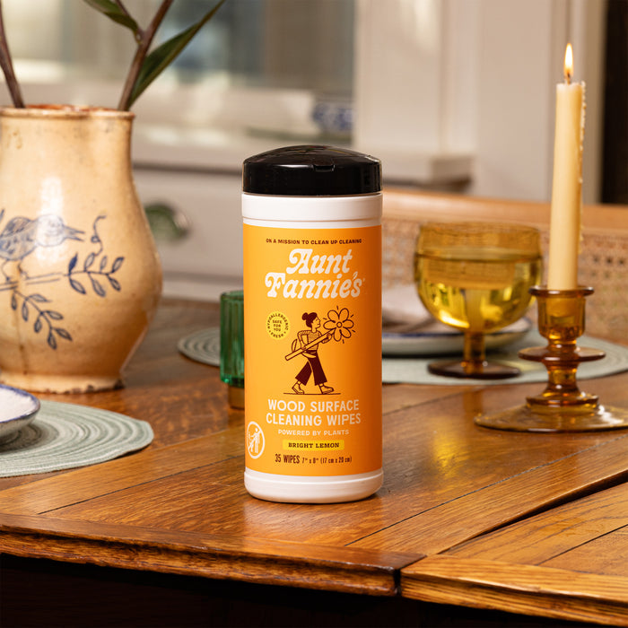 Aunt Fannies Wood Surface Wipes rest on a wooden table surrounded by a lit candle, glasses, and a ceramic vase. The label shows a woman cleaning and emphasizes the Bright Lemon scent from the natural formula with essential oils.