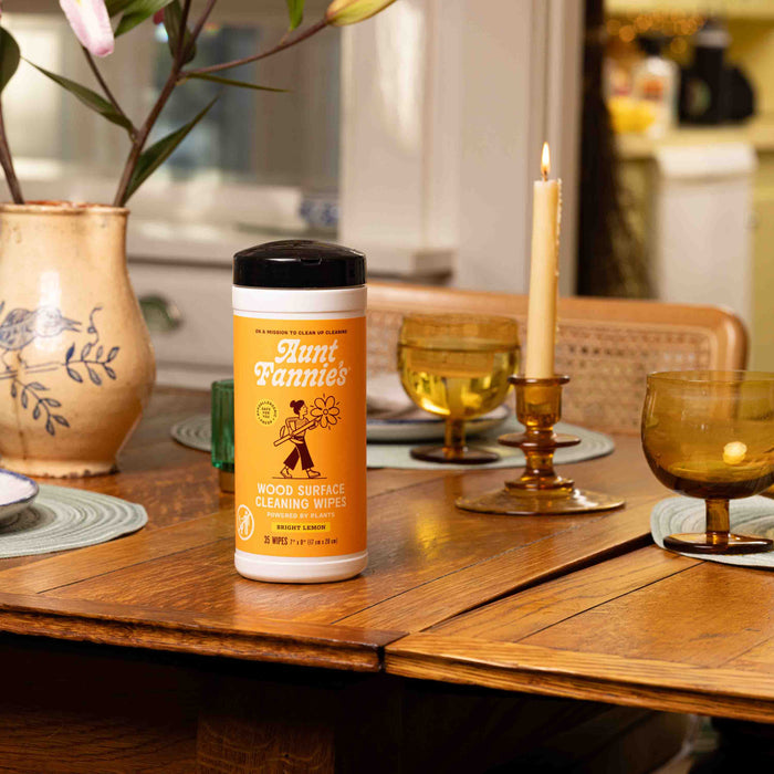 Aunt Fannies Wood Surface Wipes rest on a wooden dining table with placemats, vintage glassware, flowers in a ceramic vase, and a lit candle, creating a cozy dining room scene.