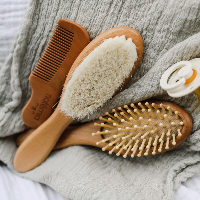 The Natemia Wooden Baby Hair Brush & Comb Set includes a plush scalp brush, a plastic pin brush, and a fine-tooth comb on a textured green cloth. Nearby is a cradle cap brush and part of a bottle.