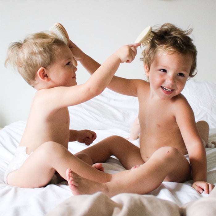 Two young children, laughing and playing on a bed with white sheets, use the Natemia Wooden Baby Hair Brush & Comb Set. One child gently brushes the others hair in a playful manner with a cradle cap brush.